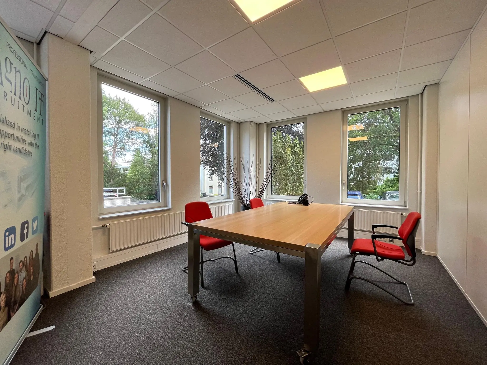 Vergaderruimte aan de Oude Utrechtseweg met houten tafel, rode stoelen en grote ramen met uitzicht op groen.